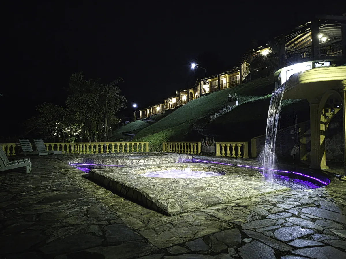 La terraza de Rancho San Antonio de noche: el jacuzzi humeando, la cascada iluminada y las habitaciones encendidas en la ladera